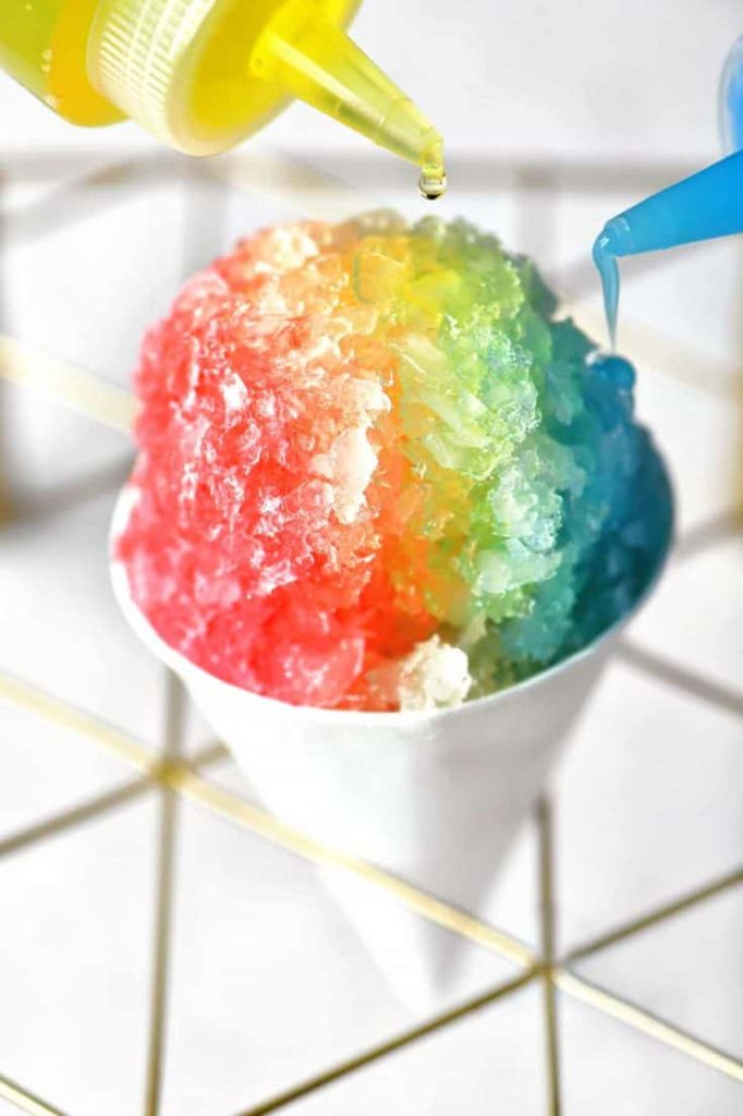 A classic rainbow snow cone in a paper cone with the syrup being poured. Une boule de neige arc-en-ciel classique dans un cône en papier avec le sirop versé