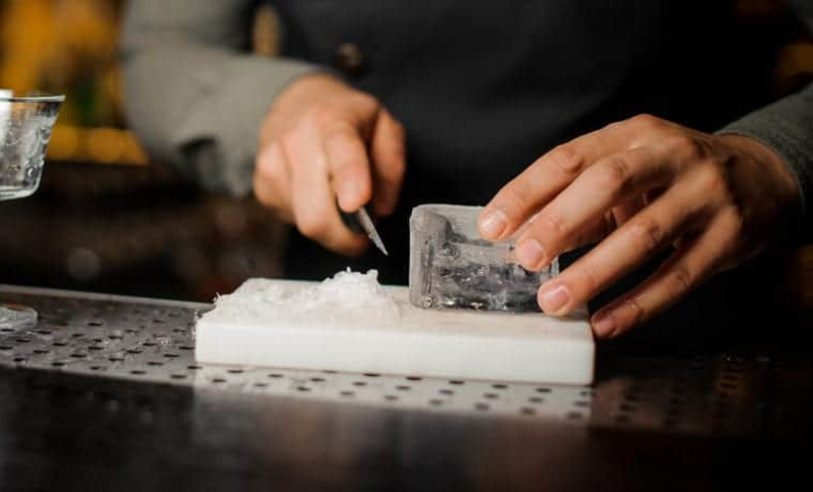 Someone shaving ice with knife on a cutting board. Quelqu’un qui rase de la glace avec un couteau sur une planche à découper.